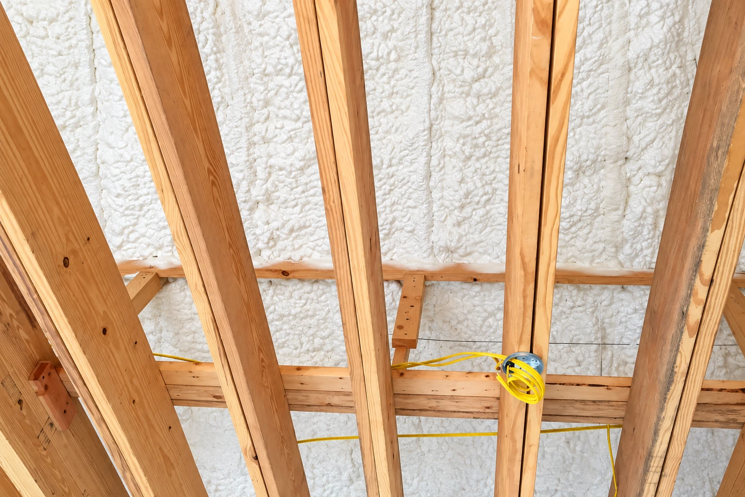 White spray foam fills the spaces between exposed wooden rafters, with a yellow electrical cable looped around one beam.