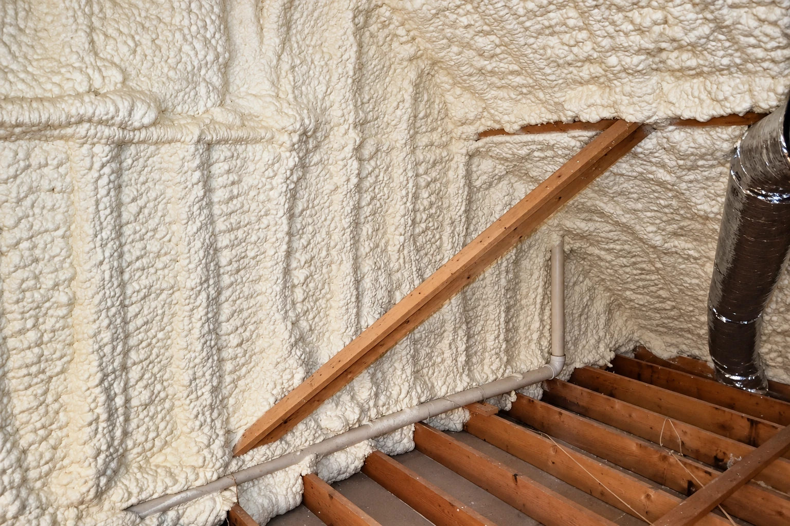 Close view of white spray foam covering a framed attic wall and roof, with exposed rafters, joists, and a pipe.