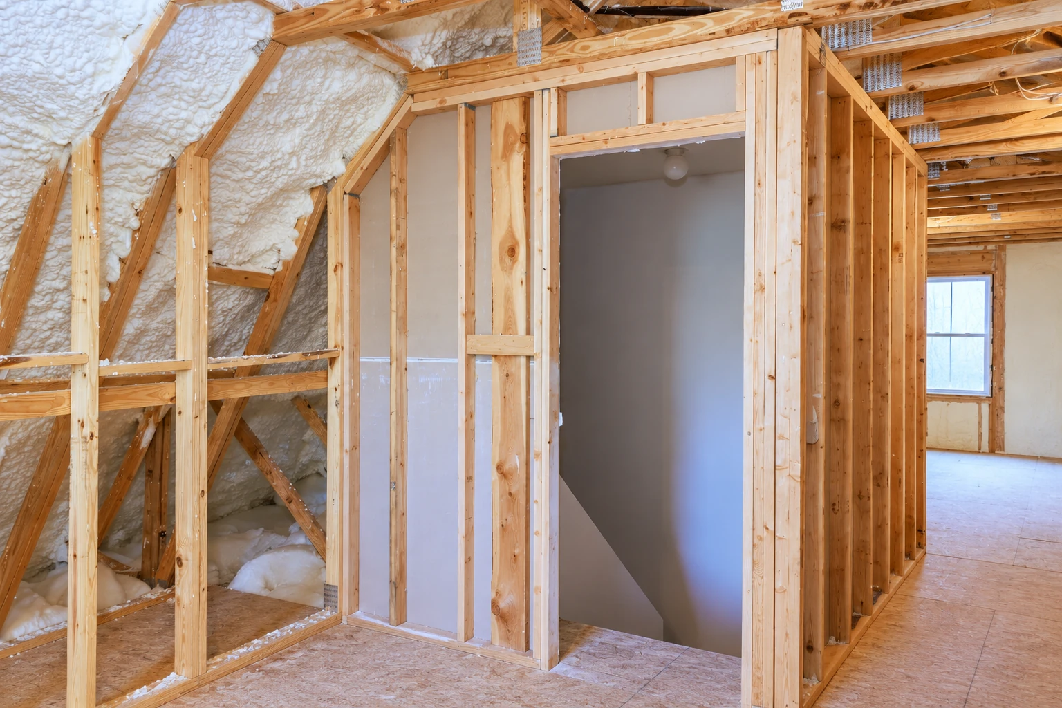 Open attic framing with spray foam insulation between the roof rafters beside an unfinished interior doorway.