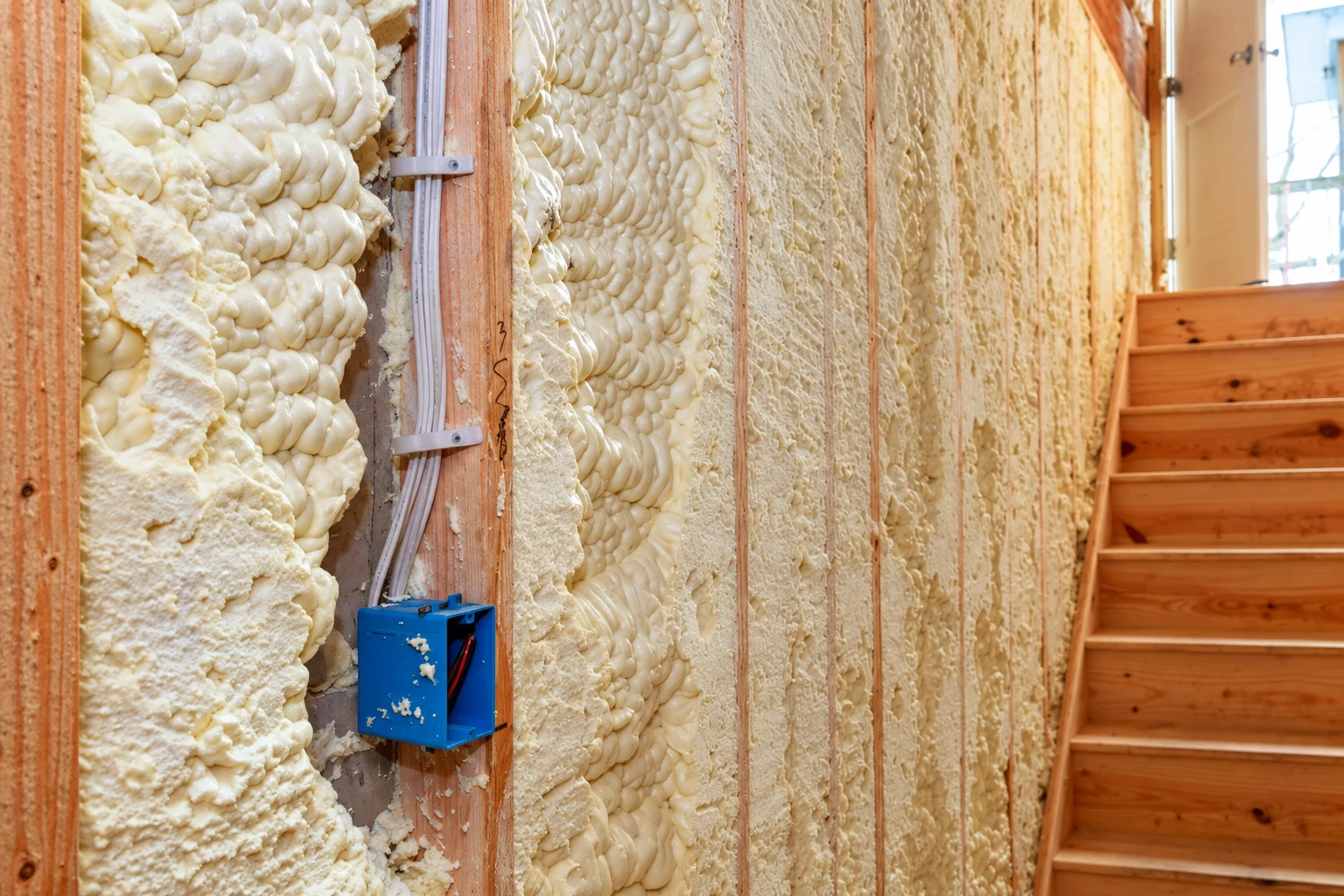 Open-cell spray foam fills exposed wall cavities between wooden studs, beside electrical wiring and an open blue electrical box.